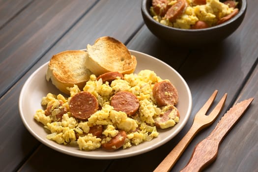 Scrambled eggs made with chorizo slices and onion on plate with toasted baguette slices, wooden fork and knife on the side, photographed with natural light (Selective Focus, Focus one third onto the plate) 