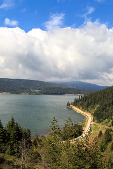 Lake or river landscape with trees and mountains around, under cloudy sky.