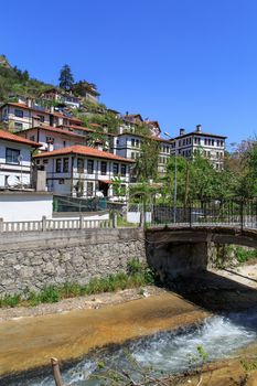 Side view of stone bridge with iron railing on river in a small town, under blue sky.