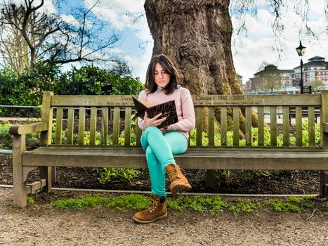 A Casual Young Woman Sitting on a Bench in a Park and Reading Her Book