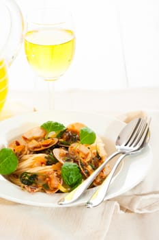 A white plate with delicious spaghetti shellfish and green basil. A glass of white wine in background.