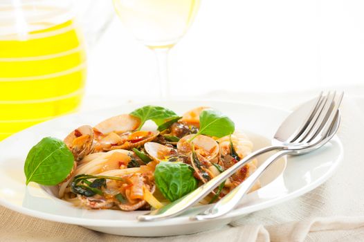 A white plate with delicious spaghetti shellfish and green basil. A glass of white wine in background.