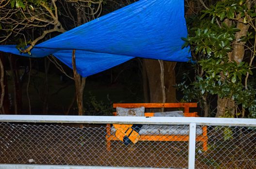 A blue tarpauling covering a bench with a mattress and cushions behind a fence in the wood