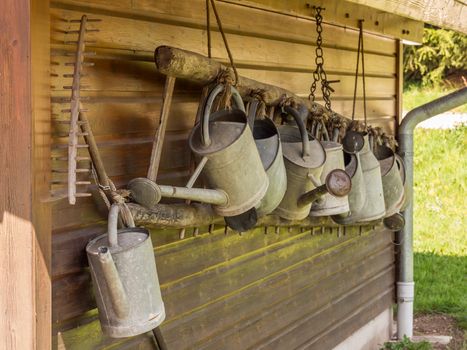A row of metal watering cans and a rake hanging in front of wooden wall