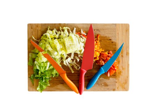 Assorted Colored Knives on Top of Wooden Cutting Board with Chopped Fresh Vegetables for Salad. Isolated on White Background.