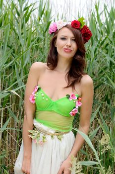 Portrait of female model with chestnut hairs, wearing wreath made of mixed flowers. Woman posing near lake surrounded by reeds.  