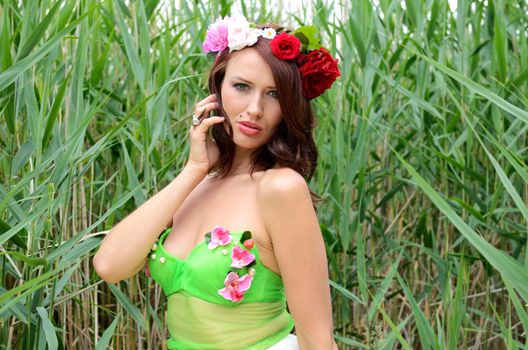 Portrait of female model with chestnut hairs, wearing wreath made of mixed flowers. Woman posing near lake surrounded by reeds.  