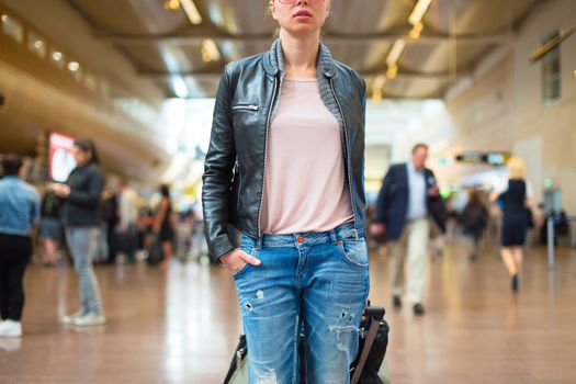 Casually dressed young stylish female traveller walking the airport terminal hall   draging suitcase behind her. Blured background. Can also be used as railway, metro, bus station.