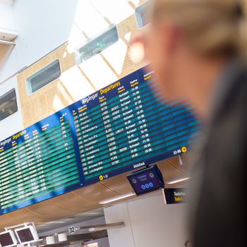 Casually dressed young stylish female traveller checking a departures board at the airport terminal hall in front of check in couters. Focus on flight schedule display.