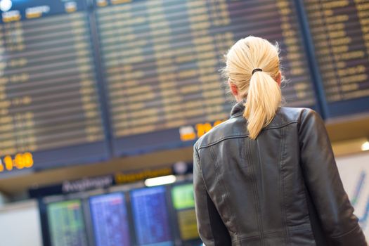 Casually dressed young stylish female traveller checking a departures board at the airport terminal hall in front of check in couters. Flight schedule display blured in the background. Focus on woman.