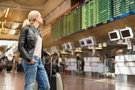 Casually dressed young stylish female traveller checking a departures board at the airport terminal hall in front of check in couters. Flight schedule display blured in the background. Focus on woman.