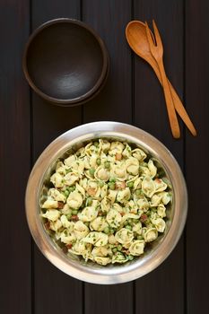 Tortellini salad with green peas, fried bacon and parsley in big salad bowl, with two small rustic bowls, wooden spoon and fork above, photographed overhead on dark wood with natural light