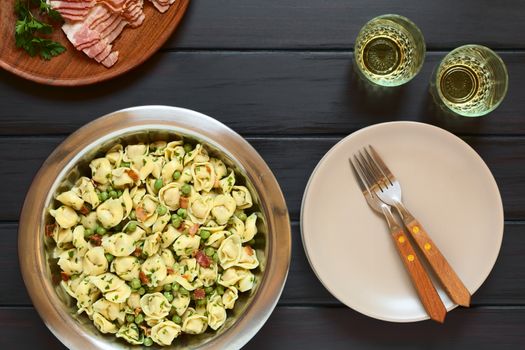 Tortellini salad with green peas, fried bacon and parsley in big salad bowl, with plates, forks and two glasses of white wine on the side, photographed overhead on dark wood with natural light