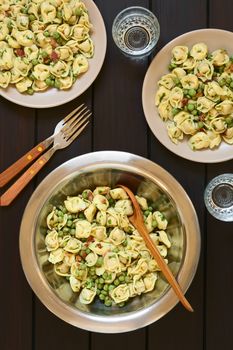 Tortellini salad with green peas, fried bacon and parsley in big salad bowl with spoon to serve, salad served on two plates, forks and glasses, photographed overhead on dark wood with natural light