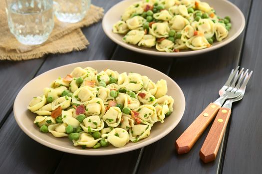 Tortellini salad with green peas, fried bacon and parsley served on plates, with forks and glasses of water on the side, photographed on dark wood with natural light (Selective Focus, Focus one third into the first salad)