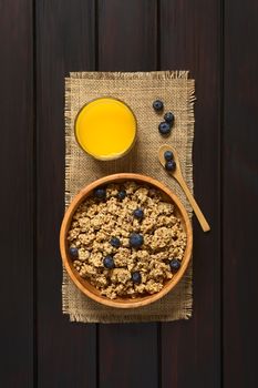 Dried berry and oatmeal breakfast cereal with fresh blueberries in wooden bowl with a glass of juice, photographed overhead on dark wood with natural light 