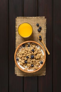 Dried berry and oatmeal breakfast cereal with fresh blueberries and milk in wooden bowl with a glass of juice, photographed overhead on dark wood with natural light 