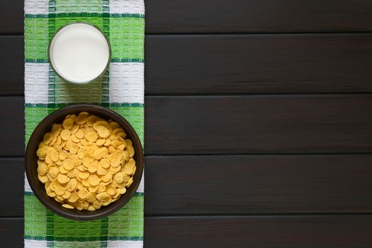 Crispy corn flakes breakfast cereal in rustic bowl with a glass of milk, photographed overhead on dark wood with natural light
