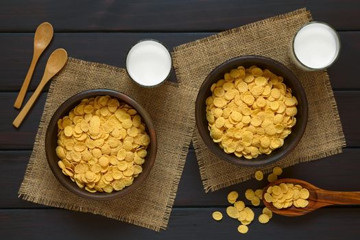 Crispy corn flakes breakfast cereal in two rustic bowls with glasses of milk and wooden spoons on the side, photographed overhead on dark wood with natural light