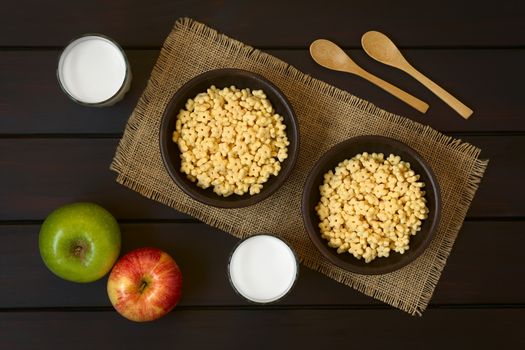 Honey flavored breakfast cereal in two rustic bowls with glasses of milk, apples and wooden spoons on the side, photographed overhead on dark wood with natural light