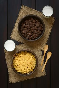 Crispy chocolate and simple corn flakes breakfast cereal in rustic bowls with glasses of milk and wooden spoons on the side, photographed overhead on dark wood with natural light