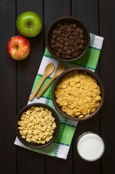 Crispy chocolate and simple corn flakes, and honey flavored breakfast cereal in rustic bowls with apples, glass of milk and wooden spoons on the side, photographed overhead on dark wood with natural light