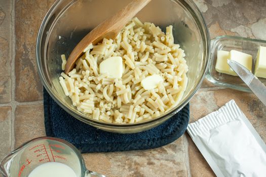 Elbow macaroni in a bowl with butter, milk nearby, and a dry cheese pack on the counter.