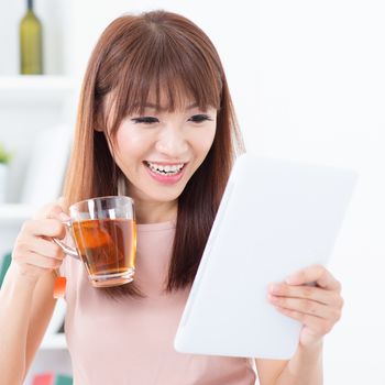 Portrait of happy Asian girl enjoying cup of hot tea while using digital pc tablet in the morning. Young woman indoors living lifestyle at home.