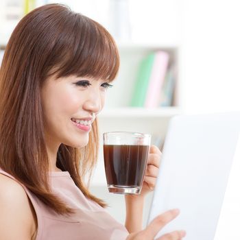 Portrait of attractive Asian girl enjoying cup of hot coffee while using computer tablet in the morning. Young woman indoors living lifestyle at home.