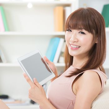 Portrait of attractive Asian girl using digital tablet and looking at camera. Young woman indoors living lifestyle at home.