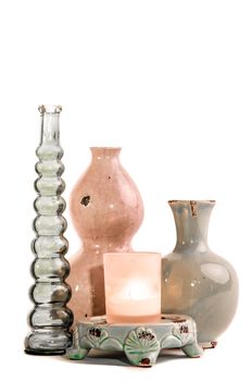 Two ceramic vases and one glass vase together with a glass holder with burning candle on small ceramic pedastil set on white background.