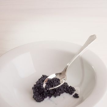 A silver spoon holds some black caviar on top of a heap of caviar inside a white bowl placed on a white wooden surface.