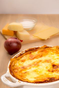 A bowl of lasagna stand in the foreground on a wooden table, while in the background some of the ingredients used, to create the dish, is visible.