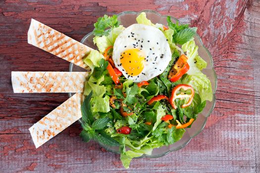 Fresh leafy green mixed salad with lettuce and herbs, tomato and sweet bell peppers served with a seasoned fried egg and wafers, viewed from above on a rustic wooden table