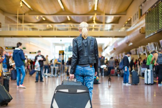 Casually dressed young stylish female traveller walking the airport terminal hall   draging suitcase and a handbag behind her. Blured background. Can also be used as railway, metro, bus station.