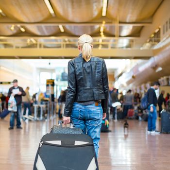 Casually dressed young stylish female traveller walking the airport terminal hall   draging suitcase and a handbag behind her. Blured background. Can also be used as railway, metro, bus station.