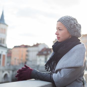 Thoughtful woman wearing winter coat, sknitted scarf, cap, and gloves standing outside on cold winter day. Beautiful romantic medieval city of Ljubljana in the background.