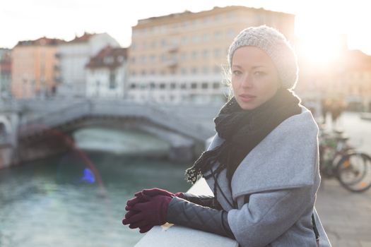 Thoughtful woman wearing winter coat, sknitted scarf, cap, and gloves standing outside on cold winter day. Beautiful romantic medieval city of Ljubljana in the background.