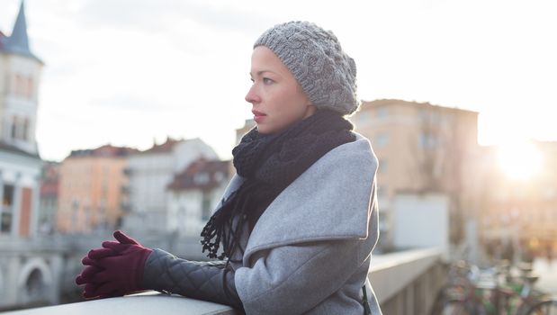 Thoughtful woman wearing winter coat, sknitted scarf, cap, and gloves standing outside on cold winter day. Beautiful romantic medieval city of Ljubljana in the background.