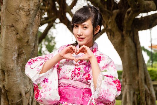 Asian woman in kimono by trees in garden, making a heart with her hands