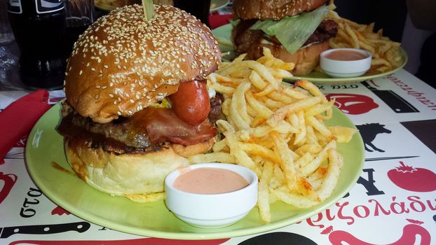 View of a huge hamburger with burger, fried eggs, sausage, barbeque sauce and bacon accompanied by fried potatoes and sauce.