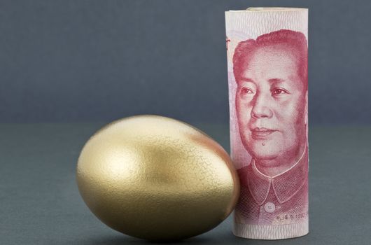Chinese currency, Asian yuan, stands upright next to gold nest egg on soothing dark background reflects Pacific Rim strength and success in business and investment.