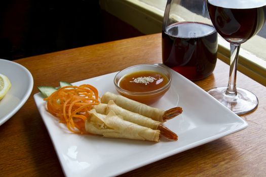 Fried prawns wrapped in pastry skin with plum sauce. Prawn presented with carrot and cucumber garnish on side on white platter.  Wine on the table.  