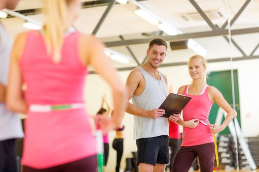 fitness, sport, training, diet, gym and lifestyle concept - two smiling people with clipboard and measure tape in the gym