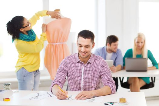 startup, education, fashion and office concept - smiling male drawing sketches and female adjusting dress on mannequin in office