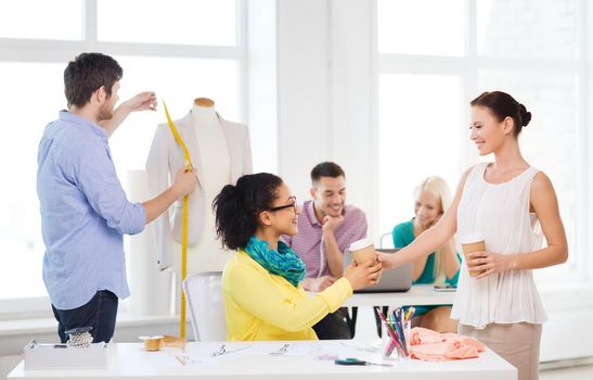 startup, education, fashion and office concept - smiling female designers having a coffee break in office with team on the back working