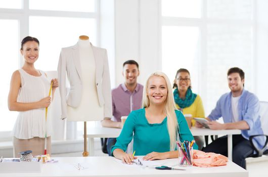 startup, education, fashion and office concept - smiling designers drawing sketches and measuring jacket on mannequin in office