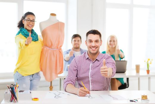 education, fashion and office concept - smiling male drawing sketches and female adjusting dress on mannequin in office and showing thumbsup