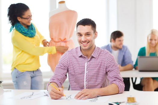 startup, education, fashion and office concept - smiling male drawing sketches and female adjusting dress on mannequin in office