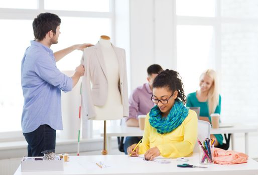 startup, education, fashion and office concept - smiling female drawing sketches and male measuring jacket on mannequin in office